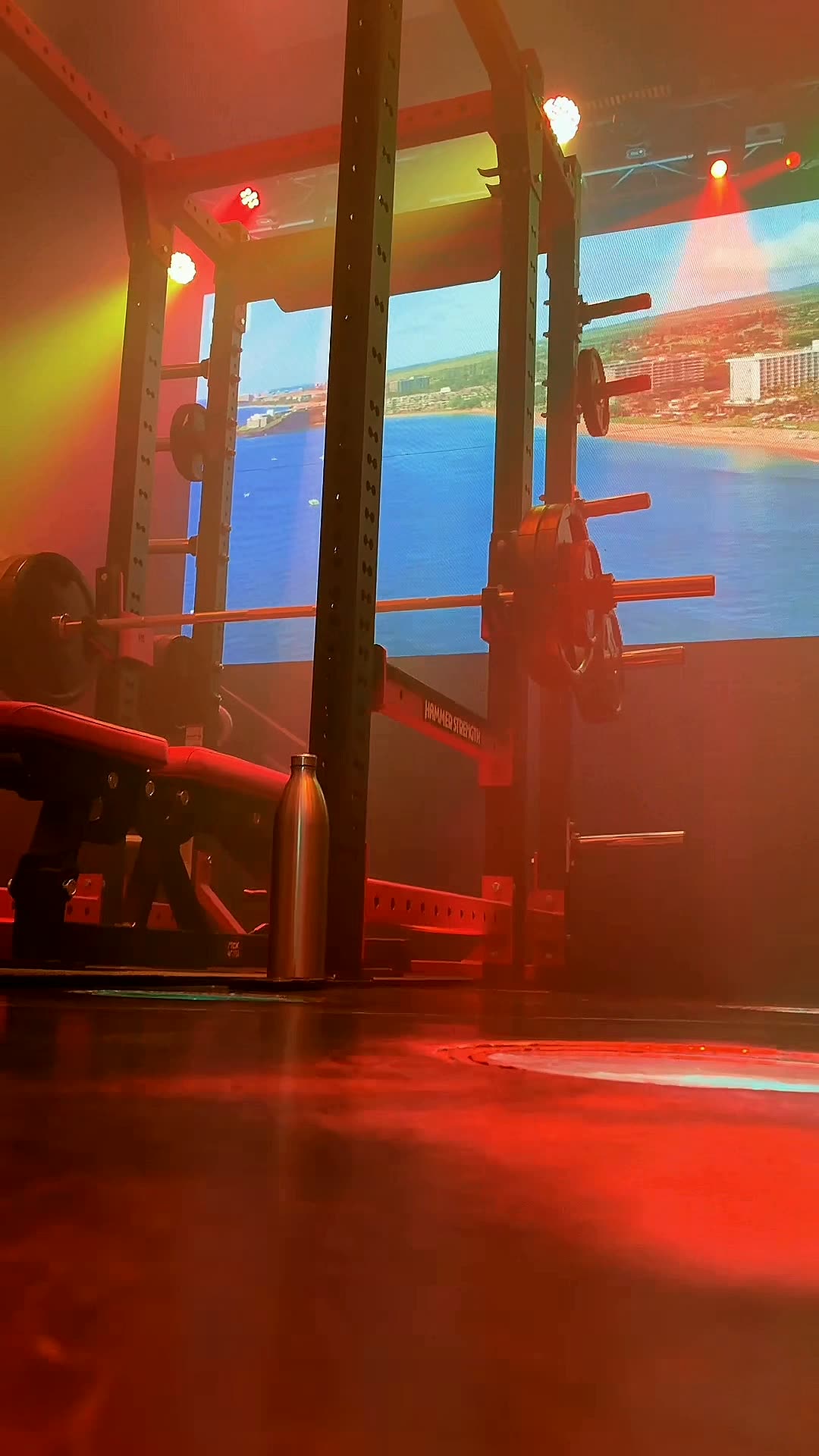 Power rack lit deep red on the training floor at Wabash Iron, with a huge LED panel showing an open coastline
